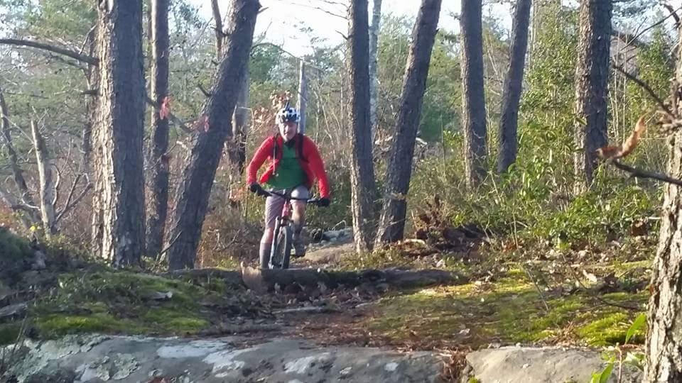 A person riding a mountain bike on a forest trail surrounded by tall trees and greenery, wearing a red jacket and helmet. Big South Fork mountain bike trail.