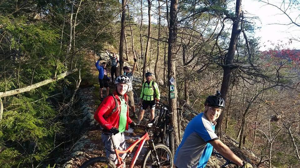 A group of six mountain bikers stands on a narrow rocky trail surrounded by trees. The riders are wearing helmets and colorful cycling attire, showcasing a mix of excitement and camaraderie. Some are holding their bikes while others pose enthusiastically for the photo, with a scenic view of wooded hills in the background. Big South Fork mountain bike trail.