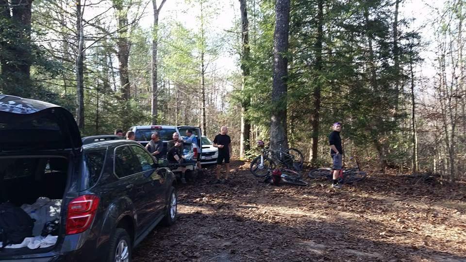Group of people gathered in a wooded area, with several bicycles leaning against trees. Vehicles are parked nearby, and leaves cover the ground, indicating an outdoor activity or gathering. Big South Fork mountain bike trail.
