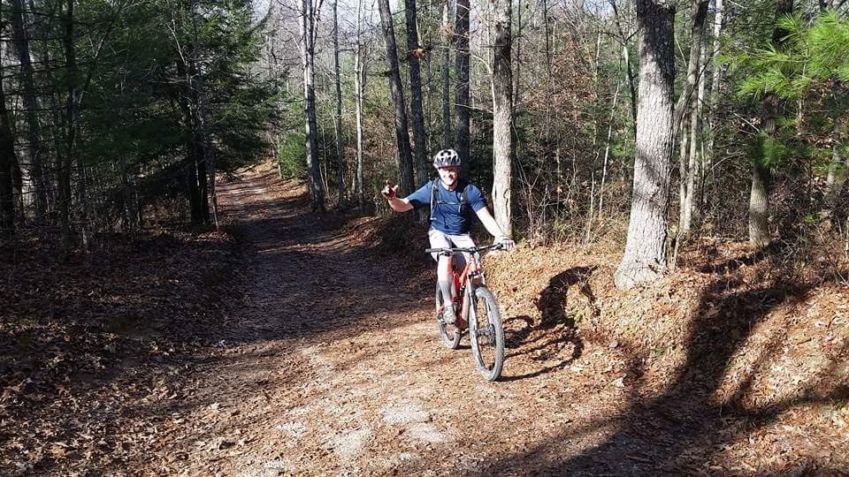 A mountain biker riding on a dirt trail through a forested area. The cyclist is wearing a helmet and appears to be waving, with trees and fallen leaves surrounding the path. Big South Fork mountain bike trail.