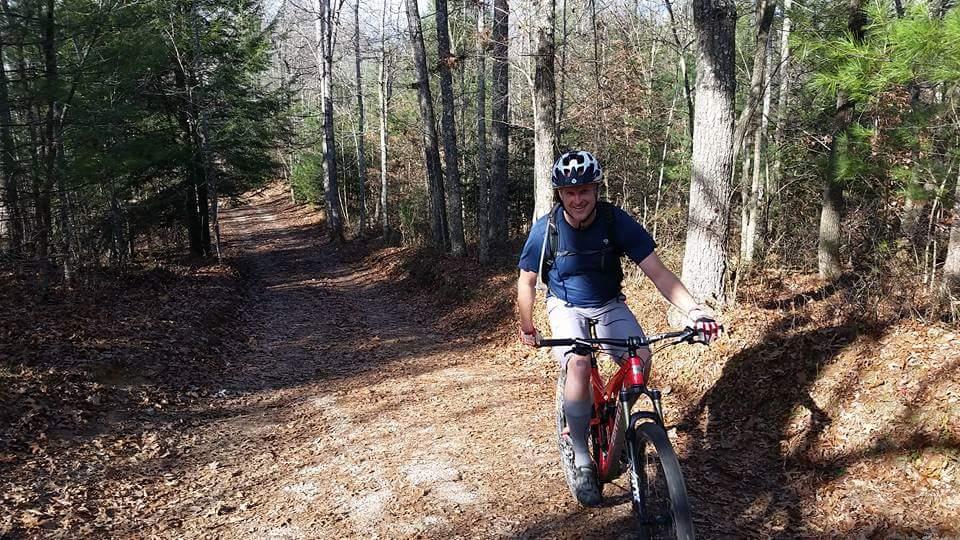 A cyclist riding a mountain bike on a dirt trail surrounded by trees. The rider is wearing a helmet and a backpack, smiling as they navigate the path lit by sunlight filtering through the foliage. The trail shows signs of leaves and natural terrain, indicating an outdoor recreational area. Big South Fork mountain bike trail.