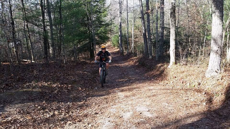 A cyclist riding a mountain bike on a dirt trail surrounded by trees in a forested area, with fallen leaves on the ground and sunlight filtering through the branches. Big South Fork mountain bike trail.