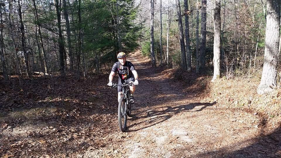 A person riding a mountain bike on a dirt trail surrounded by trees, with fallen leaves covering the ground. The scene is set in a wooded area, showcasing a sunny day with a clear blue sky. Big South Fork mountain bike trail.