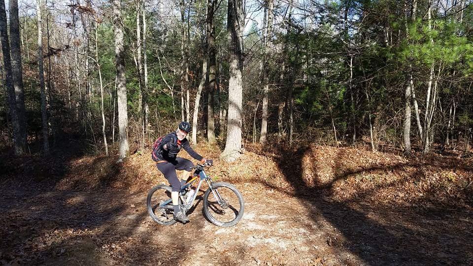 A person riding a mountain bike on a dirt trail surrounded by trees in a forest. The individual is wearing a helmet and a black jacket, with a backpack secured on their back. Sunlight filters through the trees, casting shadows on the ground covered in autumn leaves. Big South Fork mountain bike trail.