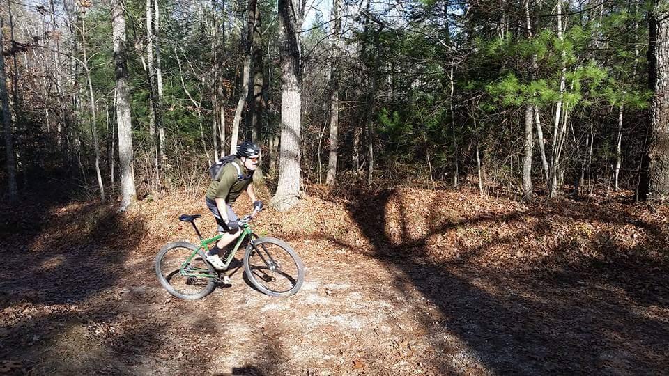 A person riding a mountain bike on a dirt trail surrounded by trees in a wooded area during autumn. The scene features a mix of sunlight and shadows on the ground, with fallen leaves and a few green plants visible in the background. Big South Fork mountain bike trail.