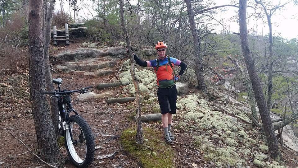A person in biking gear stands beside a mountain bike on a wooded trail, with rocky steps visible in the background. The individual is wearing a colorful jersey and helmet, leaning against a tree, with moss and foliage surrounding the scene. The atmosphere appears calm and natural, typical of a hiking or biking trail. Big South Fork mountain bike trail.