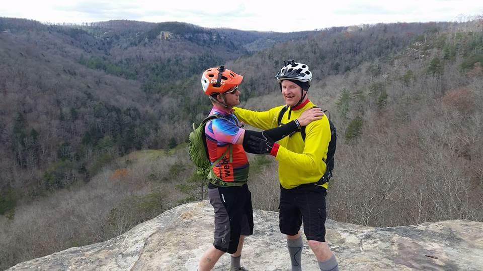Two mountain bikers stand on a rock outcrop overlooking a scenic valley filled with trees. One biker wears a colorful jersey and an orange helmet, while the other is dressed in a yellow shirt and a black helmet. They appear to be joyfully embracing each other, showcasing a moment of camaraderie against a backdrop of rolling hills and a cloudy sky. Big South Fork mountain bike trail.