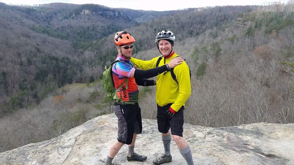 Two mountain bikers standing on the edge of a rocky outcrop, smiling and posing for the camera. They are surrounded by a scenic landscape of rolling hills and bare trees. One biker wears a colorful shirt and an orange helmet, while the other wears a bright yellow top and a black helmet. Both are dressed in cycling gear appropriate for outdoor adventures. Big South Fork mountain bike trail.