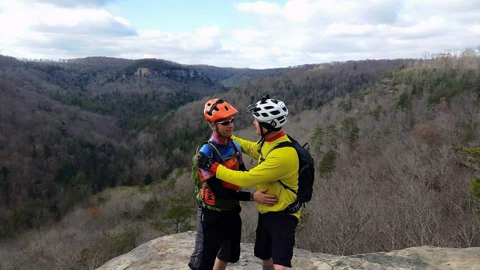 Two cyclists embrace on a rocky outcrop overlooking a lush, wooded valley. They are wearing colorful cycling gear and helmets, with a scenic backdrop of rolling hills and a partly cloudy sky. The image captures a moment of camaraderie and adventure in nature. Big South Fork mountain bike trail.