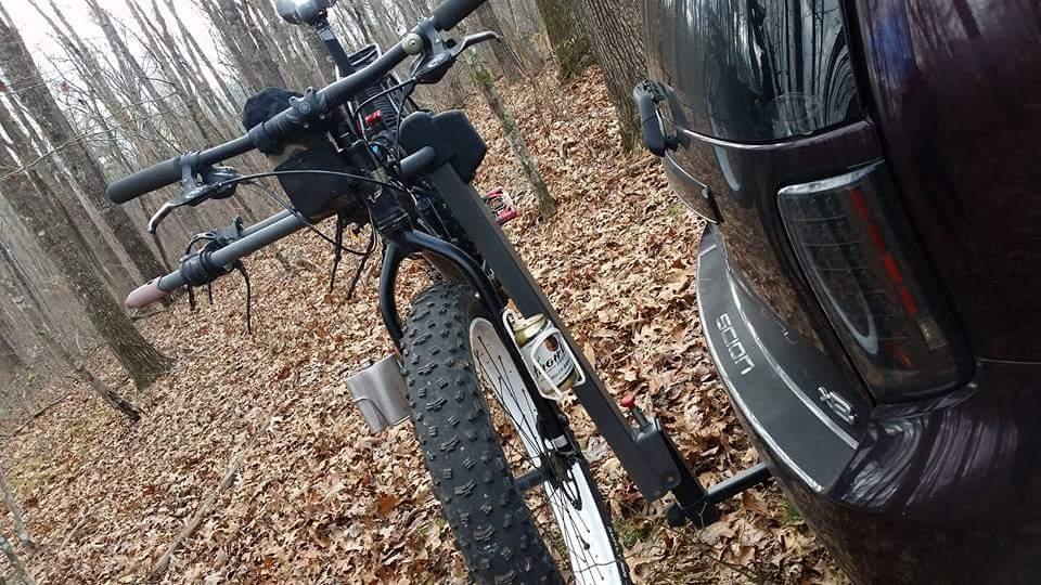 A fat tire bicycle parked next to a vehicle in a forested area, with fallen leaves covering the ground. The bike's handlebars and front tire are prominently featured, while the rear of the vehicle is visible on the right side of the image. Big South Fork mountain bike trail.
