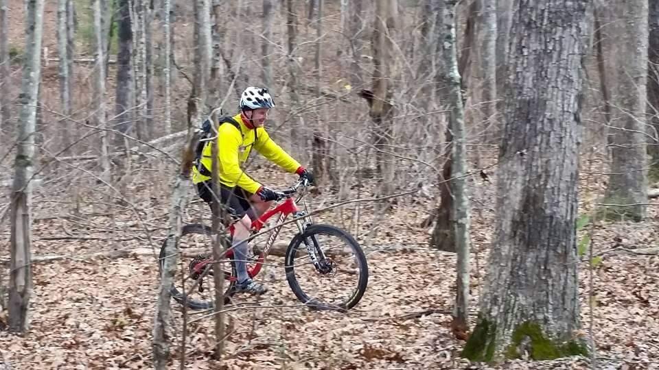 A person riding a red mountain bike through a wooded trail in a forest, wearing a yellow long-sleeve shirt and a helmet. The landscape features bare trees and fallen leaves on the ground. Big South Fork mountain bike trail.