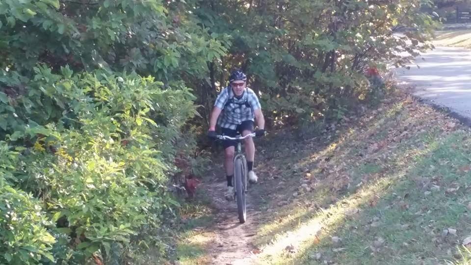 A person biking on a narrow trail surrounded by greenery, with autumn leaves scattered on the ground. The cyclist is wearing a helmet and a plaid shirt, approaching a bend in the path while sunlight filters through the trees. Brown County Park mountain bike trail.