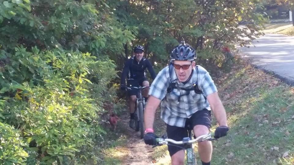 Two cyclists navigate a dirt path surrounded by lush greenery. The cyclist in the foreground, wearing a black helmet and a plaid shirt, appears focused and intense. The second cyclist, in the background, is dressed in a black outfit and is smiling, suggesting a fun outing. The setting is a sunny day, with trees providing a vibrant backdrop. Brown County Park mountain bike trail.