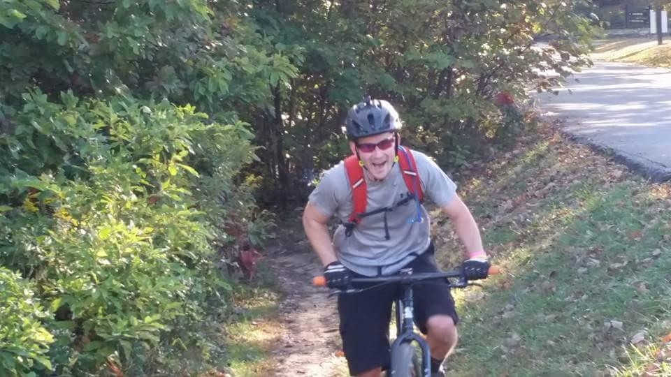 A person riding a mountain bike on a narrow trail surrounded by greenery, smiling and wearing a helmet, with a backpack and gloves. Brown County Park mountain bike trail.