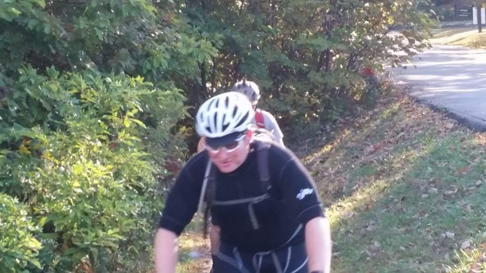 A person wearing a bike helmet and sunglasses is riding a mountain bike on a wooded path, with greenery and trees surrounding them. A second person is visible in the background, also on a bike, with a partially obscured view. The scene is set in an outdoor, natural environment, hinting at recreational biking activity. Brown County Park mountain bike trail.