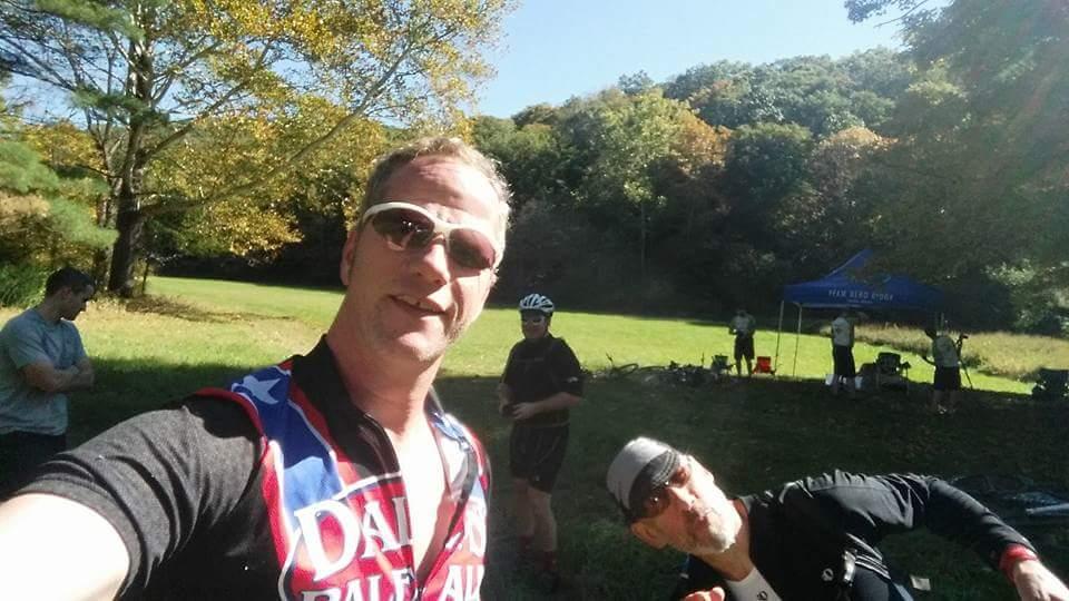 A group of cyclists posing for a selfie in a park during a sunny day. In the foreground, a person in sunglasses and a cycling jersey is smiling at the camera, while another cyclist is playfully leaning in from the side. In the background, other cyclists are gathered near a tent and enjoying the lush, autumn scenery with colorful trees. Brown County Park mountain bike trail.