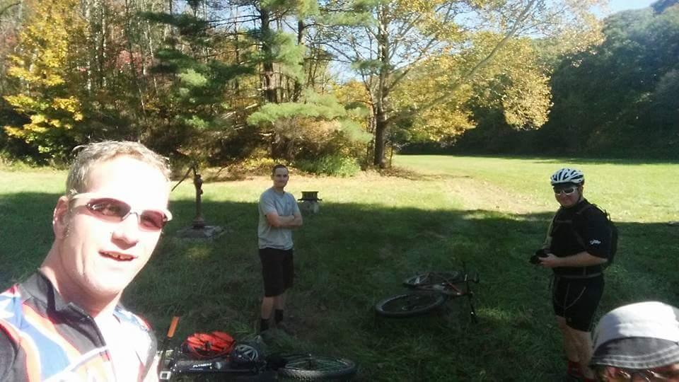 A group of three mountain bikers are taking a break in a grassy area surrounded by trees displaying autumn foliage. One cyclist is taking a selfie, while the others stand nearby, all dressed in cycling gear. Two bikes are resting on the ground beside them. Brown County Park mountain bike trail.