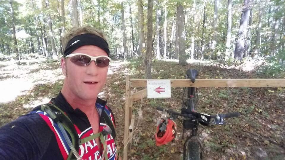 A person wearing sunglasses and a performance shirt stands in the woods next to a mountain bike. A wooden sign with an arrow points towards a trail. The background features trees with green and yellow foliage, indicating a natural outdoor setting. Brown County Park mountain bike trail.