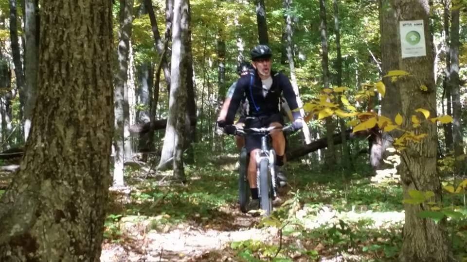 A person riding a mountain bike on a forest trail, surrounded by trees with autumn foliage. The rider is wearing a helmet and appears to be focused and determined. In the background, there is a directional sign on a tree. Brown County Park mountain bike trail.