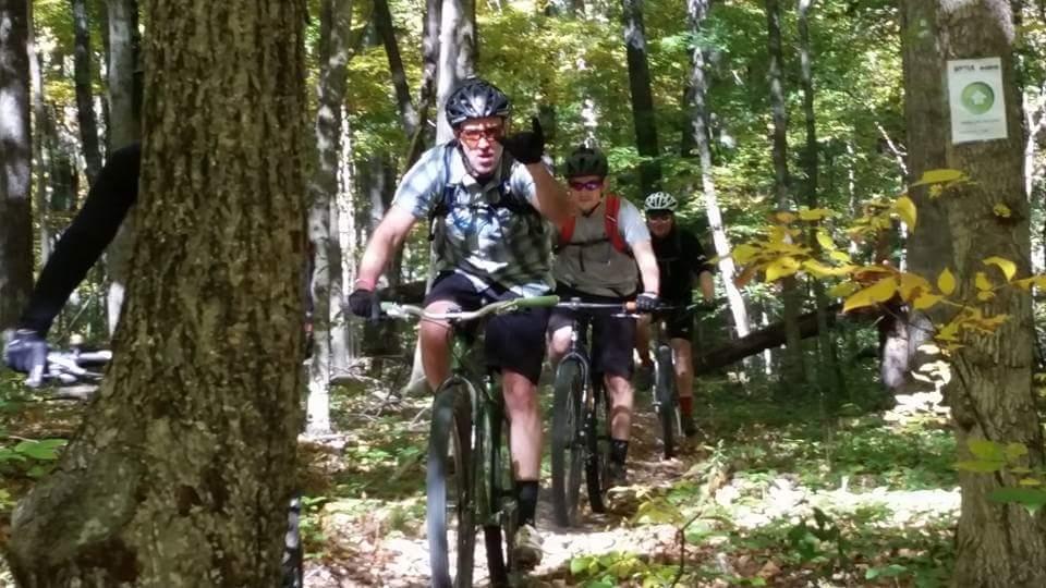 Three mountain bikers riding on a woodland trail in a sunny forest. The lead rider, wearing a helmet and a backpack, is waving to the camera. The background features trees with green leaves and autumn foliage, with a trail marker visible on a nearby tree. Brown County Park mountain bike trail.
