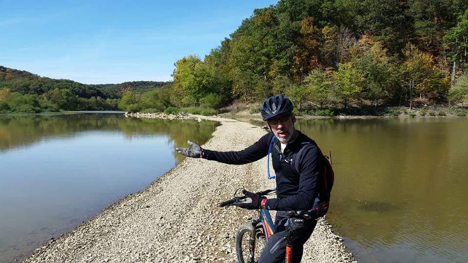 A person on a mountain bike is standing on a gravel path beside a calm river, gesturing with their hands. The background features vibrant autumn foliage on the hills under a clear blue sky. The scene conveys a sense of outdoor adventure and exploration. Brown County Park mountain bike trail.