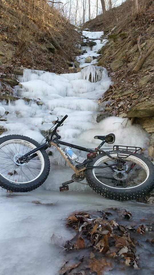A black fat bike is partially submerged in a frozen stream, surrounded by ice and scattered leaves on the ground. The icy water cascades gently down the rocky slope in the background, with bare trees lining the hillside. The scene captures the stark beauty of winter wilderness. Versailles State Park mountain bike trail.