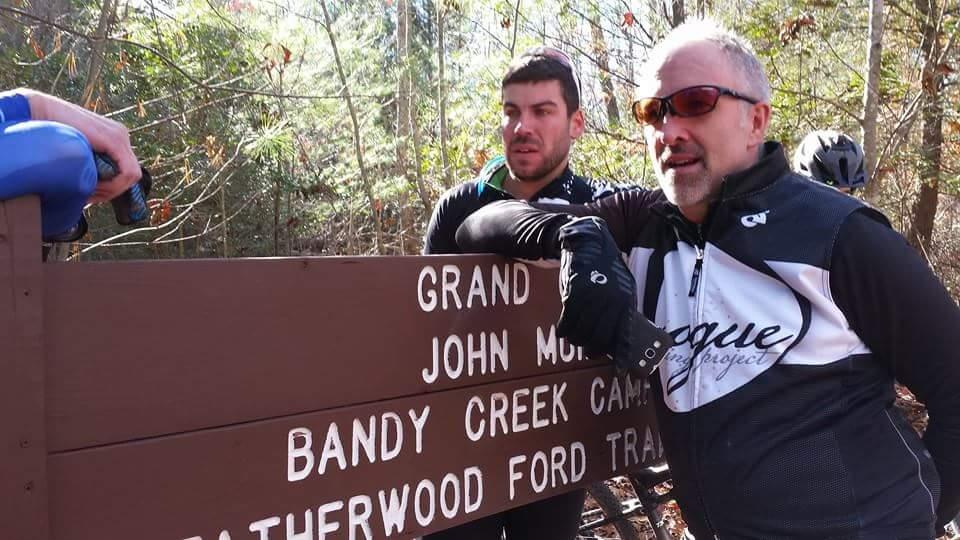 Two cyclists stand next to a wooden trail sign in a forested area, pointing out various trail names. The sign reads "Grand Junction," "John Muir," "Bandy Creek Camp," and "Heatherwood Ford Trail." One cyclist, wearing sunglasses and a black jacket, leans against the sign, while the other, in a green and black outfit, looks towards the camera. The scene is sunny with trees in the background. Big South Fork mountain bike trail.