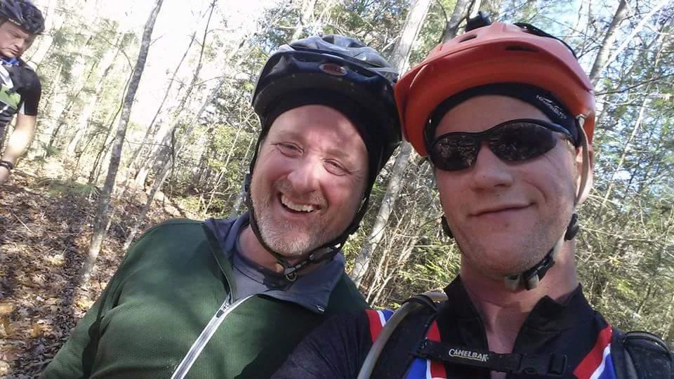 Two men smiling for a selfie while wearing helmets and biking gear in a wooded area. One man has a green jacket and the other has an orange helmet. In the background, another cyclist is partially visible. Big South Fork mountain bike trail.