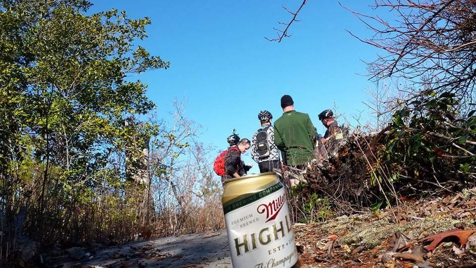 A group of cyclists gathered on a wooded trail, with a focus on a can of Miller High Life in the foreground. The background features lush greenery and a clear blue sky, creating a vibrant outdoor atmosphere. Big South Fork mountain bike trail.