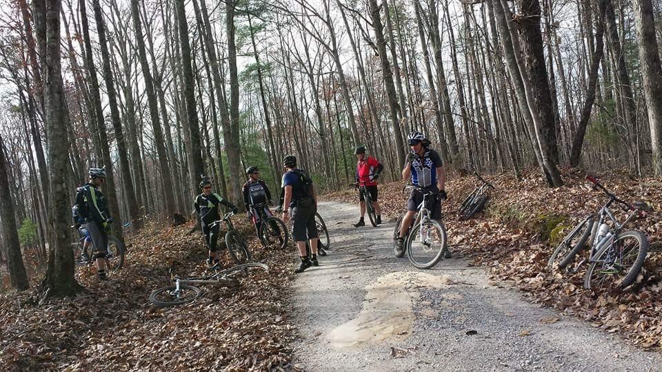 A group of six mountain bikers gathered on a gravel trail in a wooded area during autumn, surrounded by bare trees and fallen leaves. Some riders are standing with their bikes, while others are resting nearby. The scene captures a moment of camaraderie amidst nature. Big South Fork mountain bike trail.