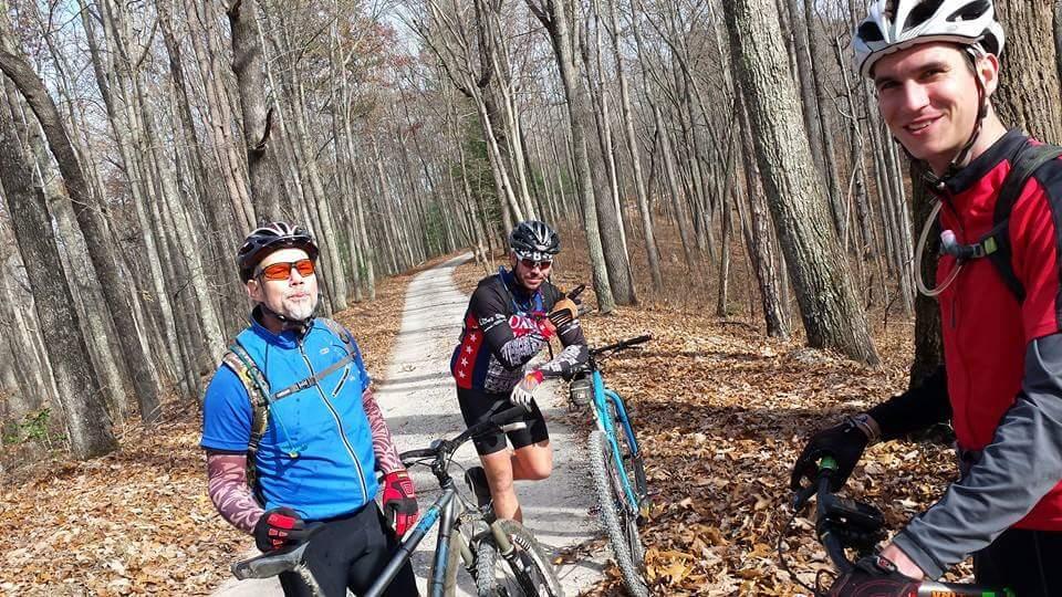 Three cyclists pause for a break on a gravel path surrounded by tall trees and autumn leaves. The cyclists are wearing colorful biking gear and helmets, and two of them are standing next to their bicycles while one is sitting on a bike. The scene captures a sunny day in a forested area. Big South Fork mountain bike trail.