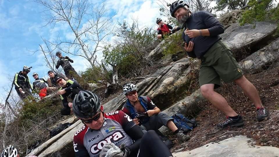 A group of cyclists resting on rocky terrain during a ride. Some are sitting on the rocks, eating or taking pictures, while others stand nearby, enjoying the outdoor scenery. The backdrop features trees and a blue sky with clouds. Big South Fork mountain bike trail.