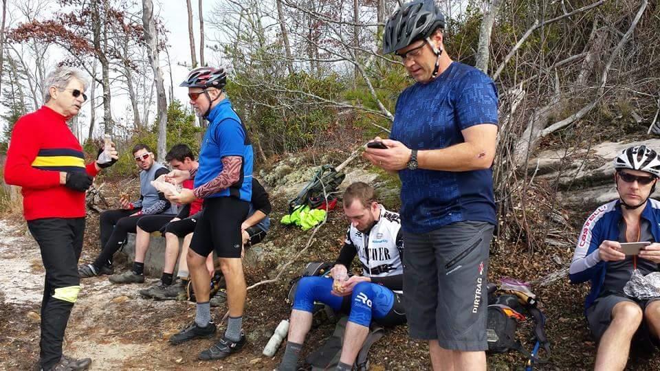 A group of seven cyclists resting on a rocky area surrounded by trees. Some are sitting on the ground, while a few are engaged in conversation, looking at their phones, or eating snacks. One cyclist is dressed in a bright red and yellow jersey, and another is wearing a blue jersey. The atmosphere appears relaxed and casual, with the cyclists taking a break during their ride. Big South Fork mountain bike trail.