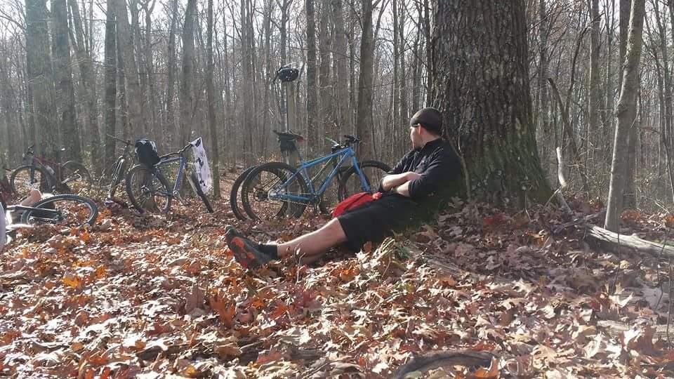 A person sitting against a tree in a forested area, surrounded by several parked bicycles. The ground is covered with autumn leaves, and the scene is illuminated by sunlight filtering through the trees. Big South Fork mountain bike trail.