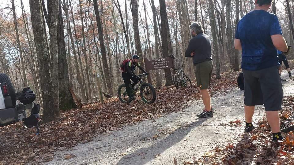 A group of mountain bikers gathered at a trailhead in a wooded area during autumn. One cyclist is adjusting their bike near a trail sign, while others are standing nearby, some with their backs to the camera. The ground is covered in fallen leaves, and trees with bare branches are visible in the background. A pickup truck is parked to the left of the image. Big South Fork mountain bike trail.