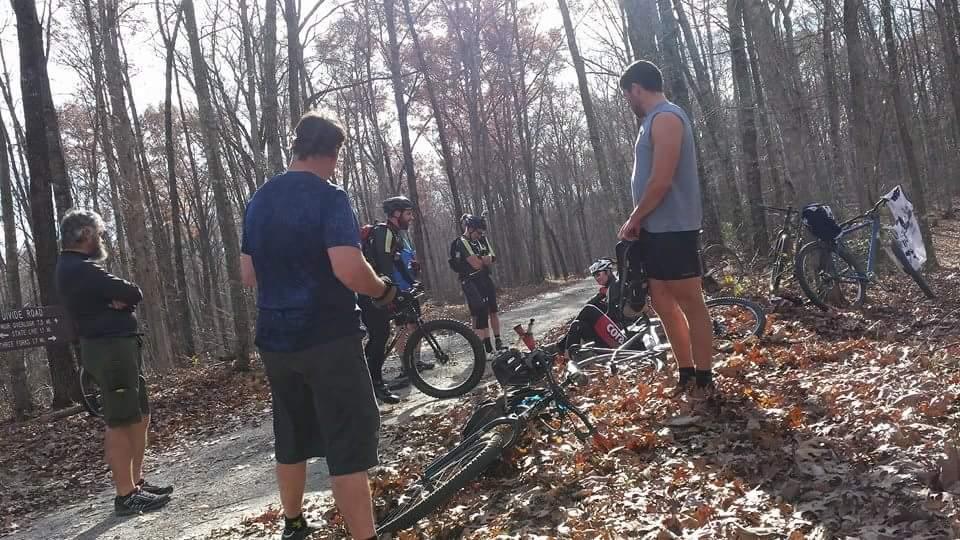 A group of five cyclists gathered on a gravel path in a wooded area, surrounded by trees with bare branches. Some bikes are leaning against the ground, and a signpost is visible in the background. The cyclists are engaged in conversation, wearing cycling attire and helmets, with fallen leaves scattered on the ground. Big South Fork mountain bike trail.