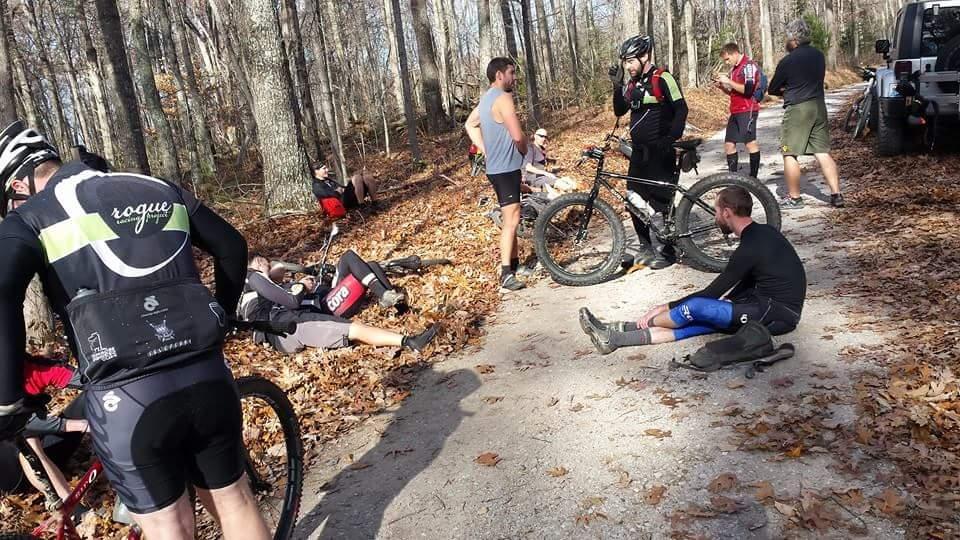 A group of mountain bikers resting on a wooded trail covered with fallen leaves. Some riders are seated or lying on the ground, while others are standing and chatting. Bicycles are scattered around, and the scene captures a relaxed moment in nature after a ride. Big South Fork mountain bike trail.