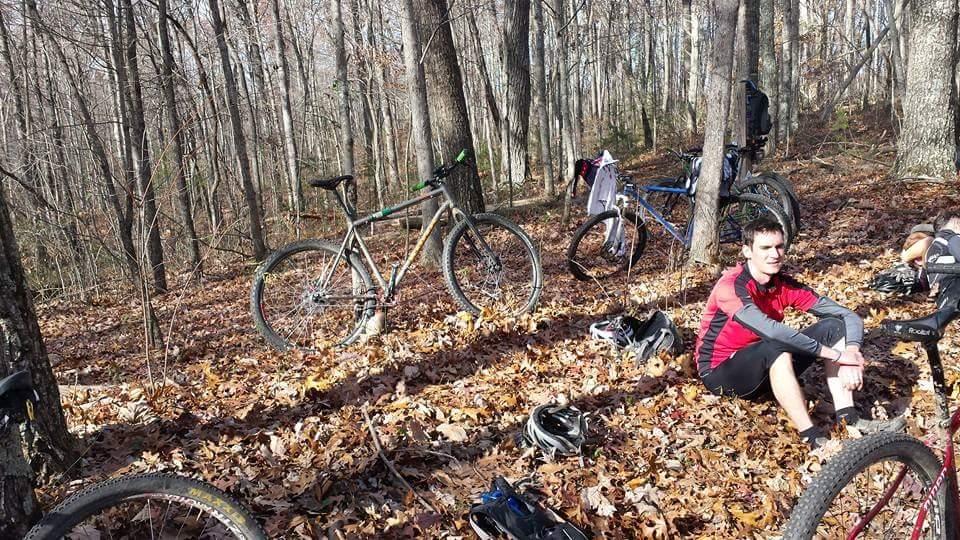 A cyclist resting on a bed of fallen leaves in a wooded area, with several mountain bikes parked nearby. The scene captures autumn foliage and a peaceful, natural environment. Big South Fork mountain bike trail.