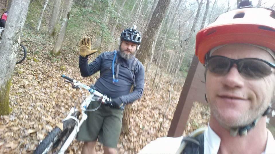 Two mountain bikers are taking a selfie on a wooded trail. One rider, wearing a white shirt and sunglasses, is in the foreground, smiling at the camera. The second rider, with a beard and wearing a black helmet and blue long-sleeve shirt, is standing beside a bike and waving. The scene is surrounded by trees and fallen leaves, indicating an outdoor setting. Big South Fork mountain bike trail.