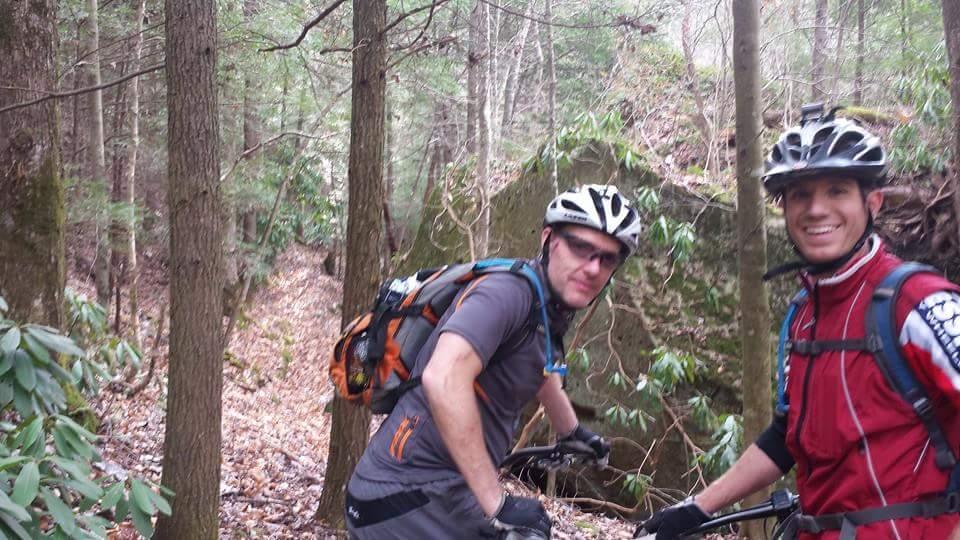 Two mountain bikers smiling and posing for the photo on a wooded trail, surrounded by trees and greenery. One biker wears a grey shirt with an orange design and an orange backpack, while the other is dressed in a red jacket. They both have helmets on and are standing near a large rock in the background. Big South Fork mountain bike trail.
