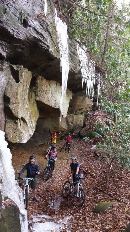 A group of mountain bikers standing on a forest trail near a rocky overhang, with icicles hanging from the cliff above. The ground is covered in fallen leaves, and there are lush green plants surrounding the scene. The bikers are smiling and enjoying their outdoor adventure. Big South Fork mountain bike trail.