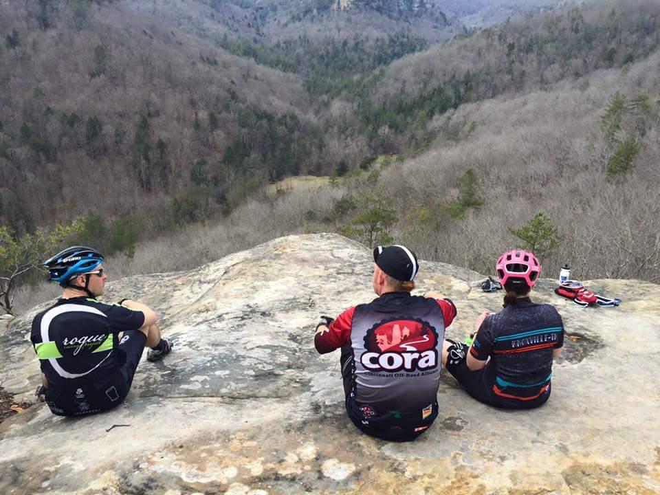 Three cyclists resting on a rocky outcrop, overlooking a valley filled with trees in early spring. The cyclists are dressed in colorful biking gear, with one wearing a blue helmet and two wearing caps. The landscape features a mix of rocky terrain and wooded hills in the background under a cloudy sky. Big South Fork mountain bike trail.