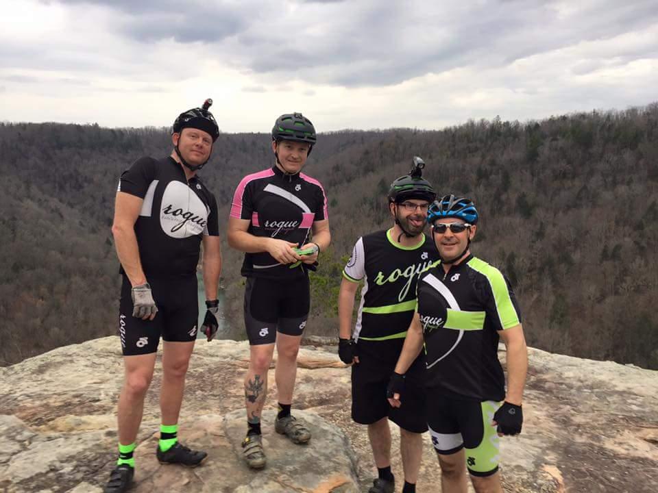 Four cyclists posing on a rocky outcrop with a scenic forested landscape in the background. They are wearing matching black and colorful cycling jerseys, helmets, and gloves, smiling and enjoying their outdoor adventure. The sky is partly cloudy, and the area appears to be a natural riding trail. Big South Fork mountain bike trail.