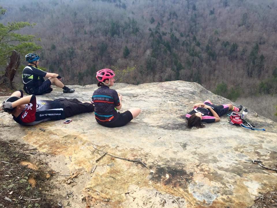 A group of four mountain bikers resting on a rocky ledge, overlooking a wooded valley. Two individuals are lying down on the rock surface, while the other two are seated, enjoying the scenery. The riders are wearing cycling gear and helmets, surrounded by a natural landscape of trees and hills in the background. Big South Fork mountain bike trail.