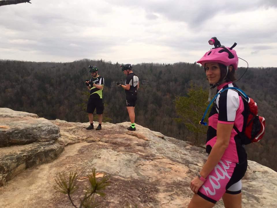 Three cyclists stand on a rocky outcrop overlooking a tree-filled valley under a cloudy sky. The first cyclist, wearing a pink helmet and a pink and black cycling outfit, looks back at the camera with a smile. The two others, dressed in cycling gear, appear to be checking their devices. The landscape is dotted with trees, indicating a natural setting perfect for outdoor activities. Big South Fork mountain bike trail.