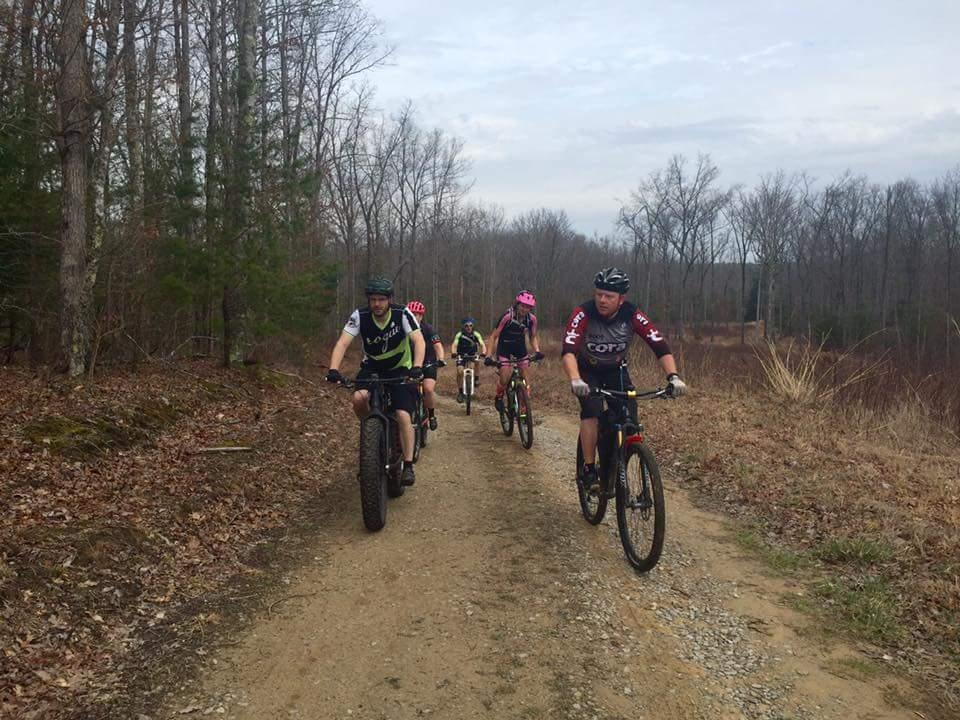 A group of five cyclists riding on a dirt trail surrounded by trees. The riders are wearing various cycling gear and helmets, with a mix of mountain bikes, including a fat tire bike. The trail is well-defined with some gravel, and the landscape shows early signs of spring with sparse leaves on the trees. The sky is overcast. Big South Fork mountain bike trail.
