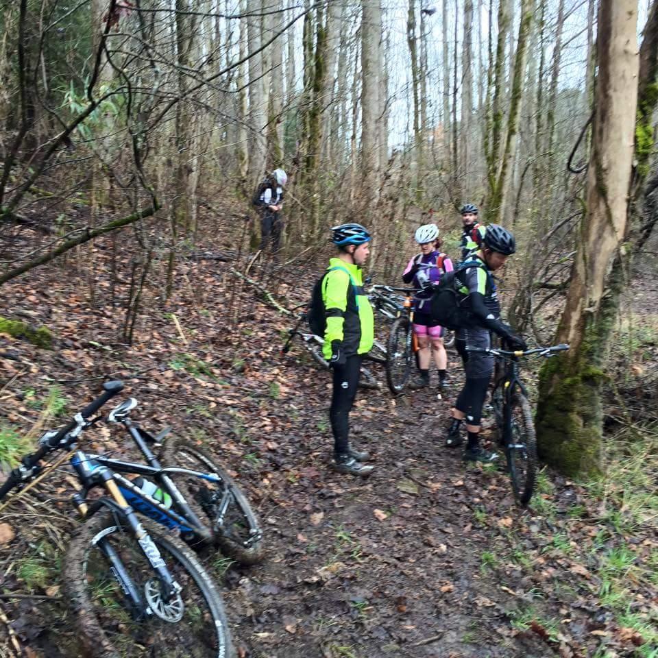 A group of mountain bikers gathered on a muddy trail in a forest, with several bicycles leaning against the ground. The scene captures the riders in various cycling gear, discussing or resting amidst tall trees and fallen leaves. Big South Fork mountain bike trail.
