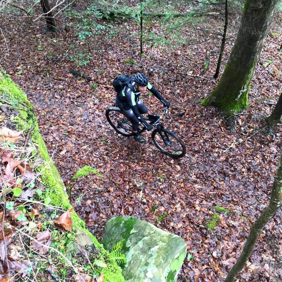 A cyclist rides a mountain bike on a trail covered in fallen leaves, surrounded by trees in a wooded area. The scene captures a dynamic perspective from above, highlighting the natural terrain and greenery. Big South Fork mountain bike trail.
