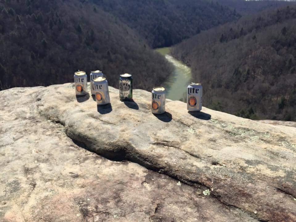 A row of six beer cans placed on a rocky cliff overlooking a river and wooded valley. The sun is shining, creating a bright, outdoor scene. Big South Fork mountain bike trail.