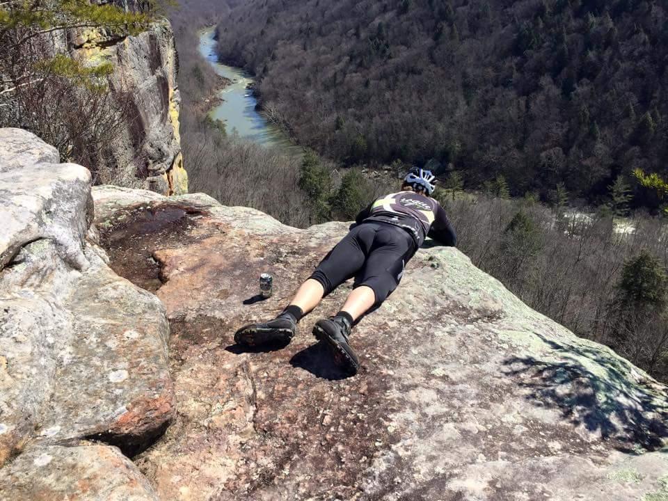A cyclist lying on a rocky outcrop, looking over a steep cliff into a valley below, with a river winding through the landscape. The surrounding area features bare trees, indicating early spring, and a can is placed near the cyclist. Big South Fork mountain bike trail.